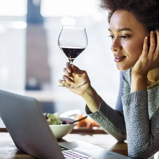 Photo of woman with a glass of wine, looking at her laptop.